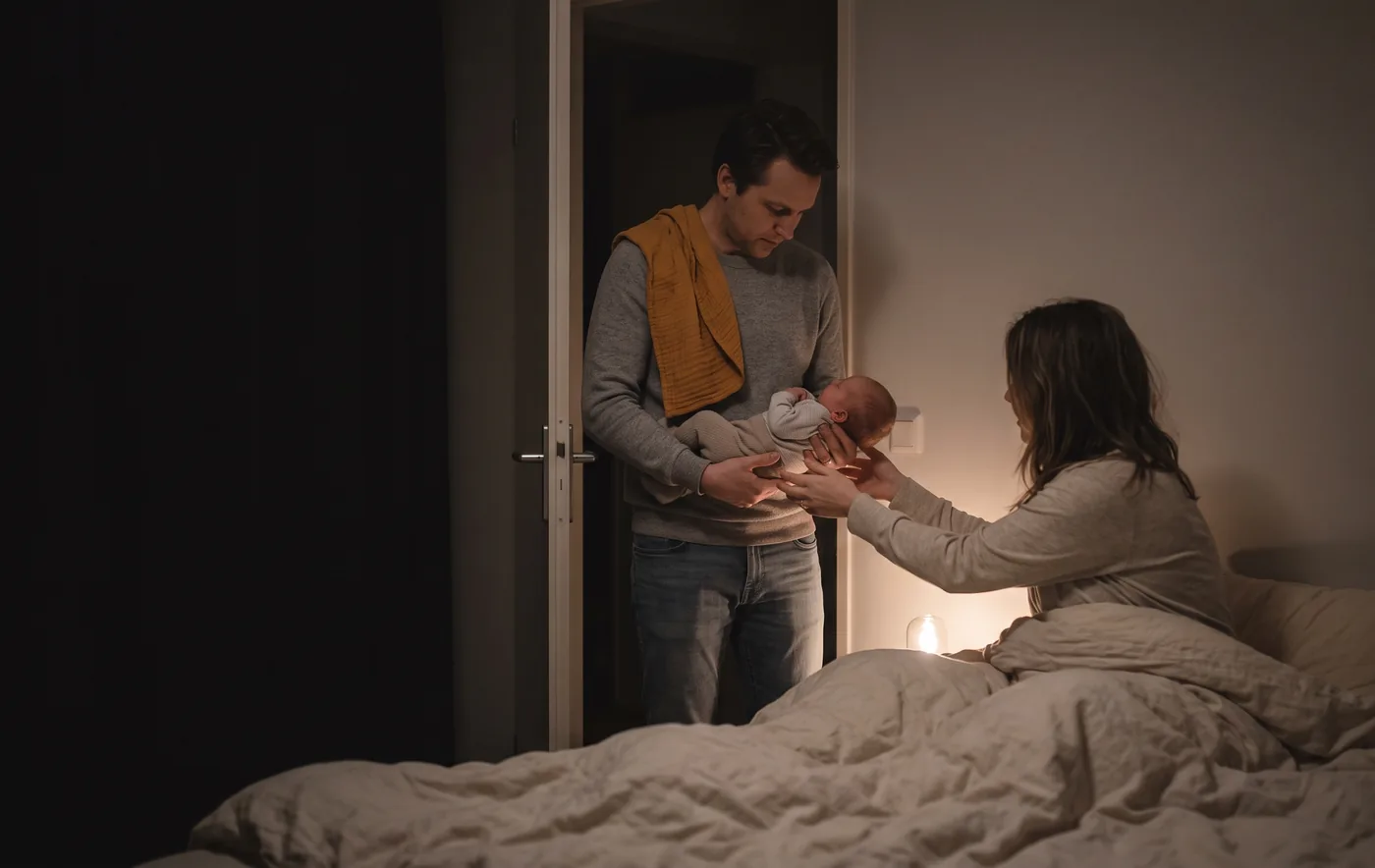Two parents passing their newborn between them at night in a dimly lit bedroom, one standing in the doorway with an ochre muslin over their shoulder, the other sitting up in bed reaching out to take the baby